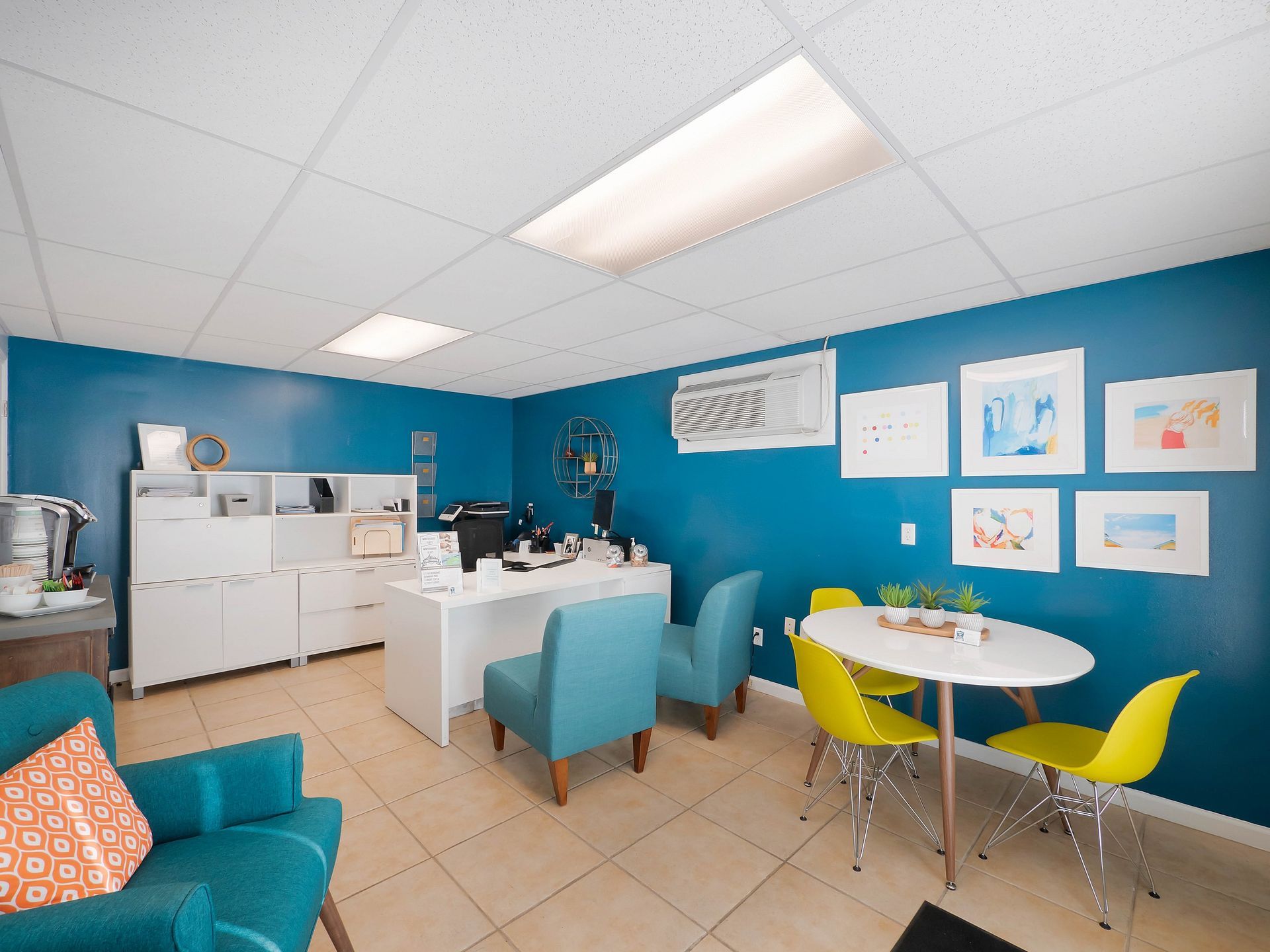 A bright office with teal walls, a white desk, two teal armchairs, and a small round table with two yellow chairs.