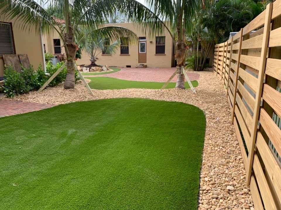 Landscaped backyard with lush green lawn, palm trees, gravel paths, and a wooden privacy fence.