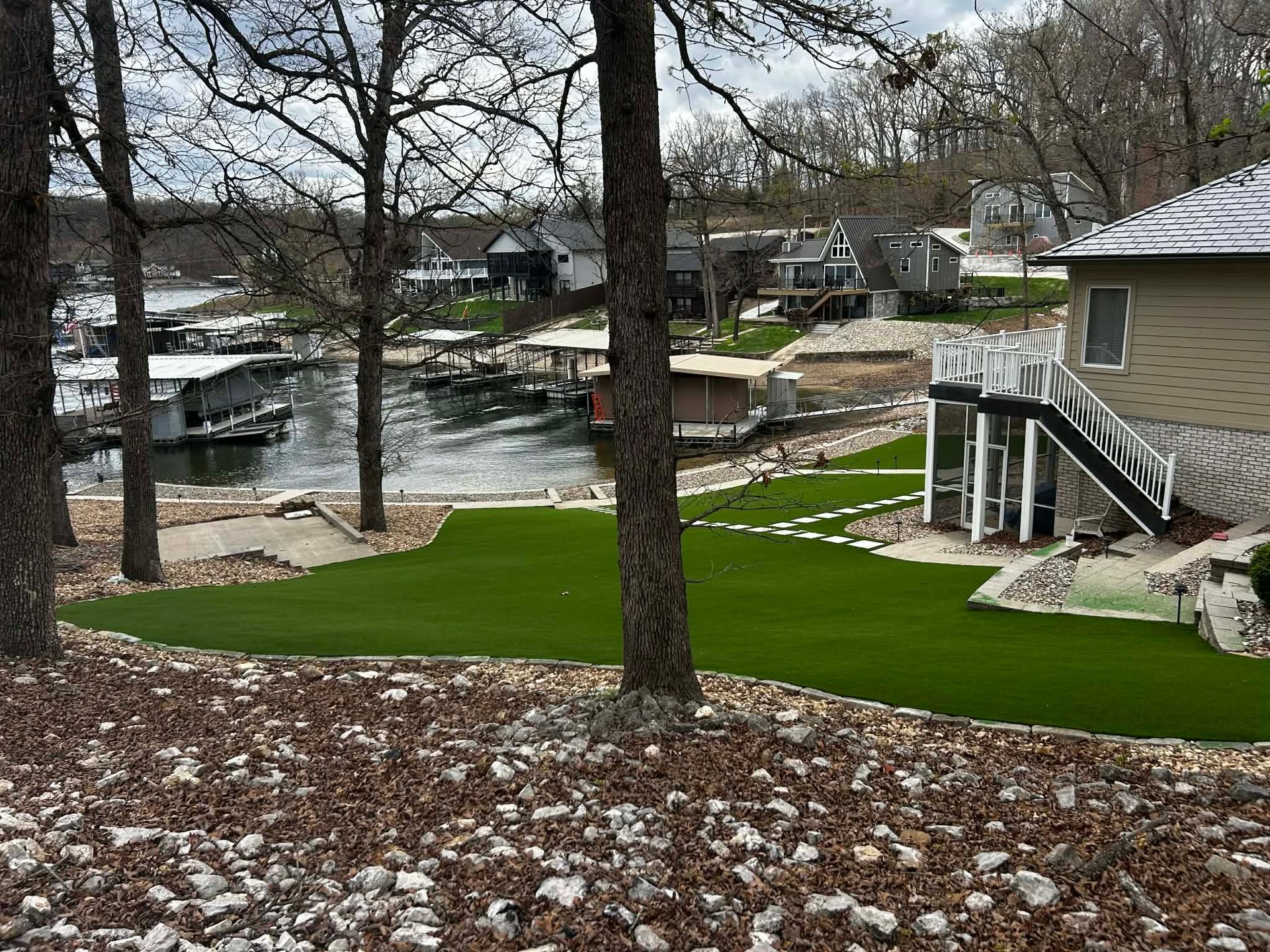 Snow-dusted yard beside a river with trees, green lawn, and a house with stairs and a patio.