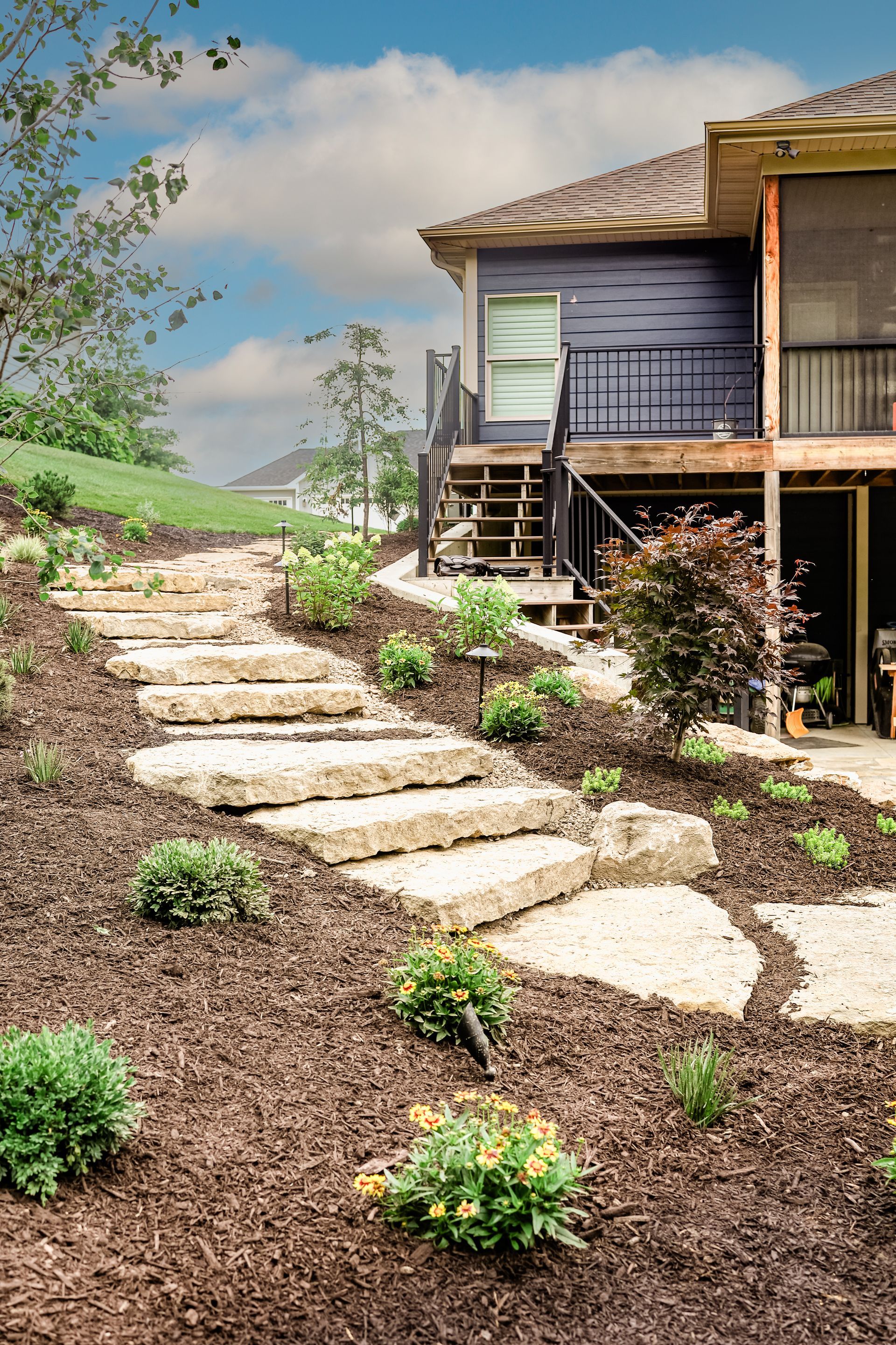 Stepping-stone path through mulch leads to a house porch with shrubs and clear sky.