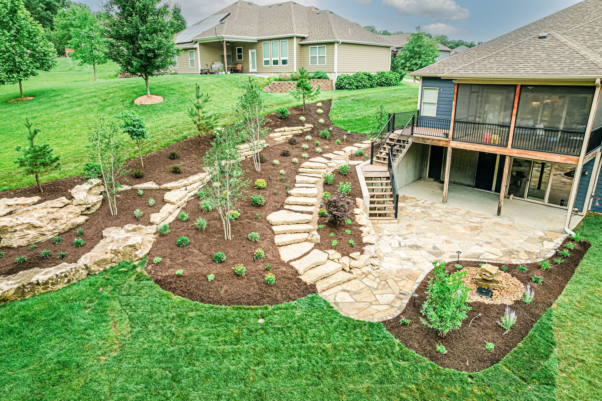 Backyard landscaping with curved stone paths, mulch beds, young plants, and a deck beside a house