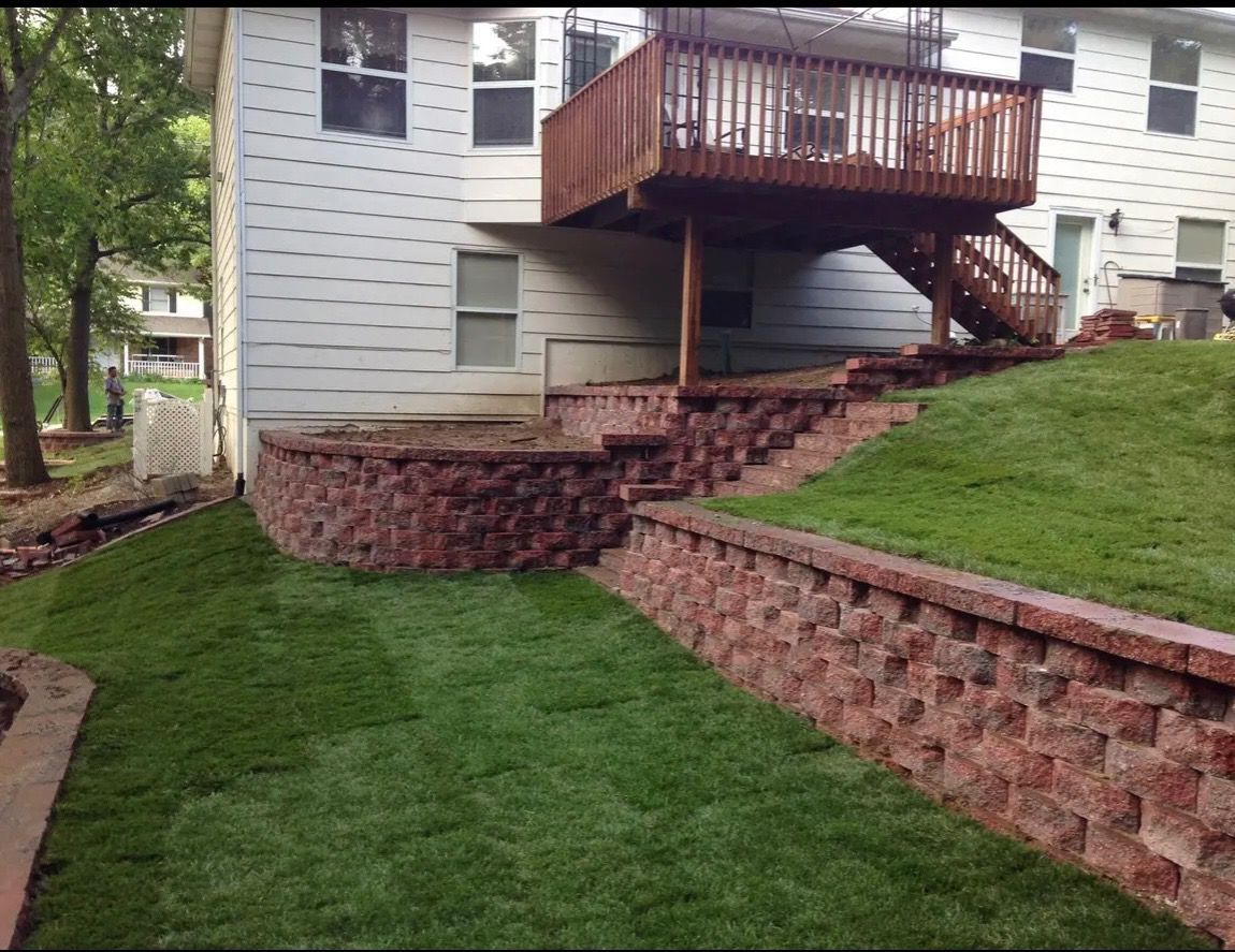House with raised wooden deck, brick retaining wall, and green sloped lawn