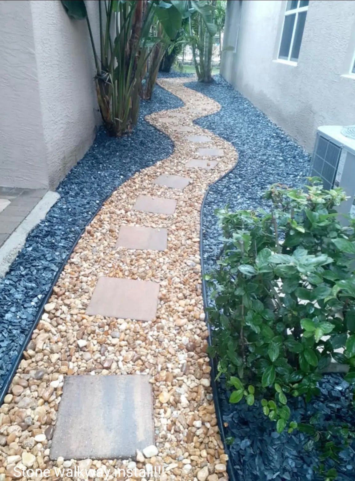 Narrow curved garden path of tan pavers and stepping stones between a house and plants, bordered by dark gravel.