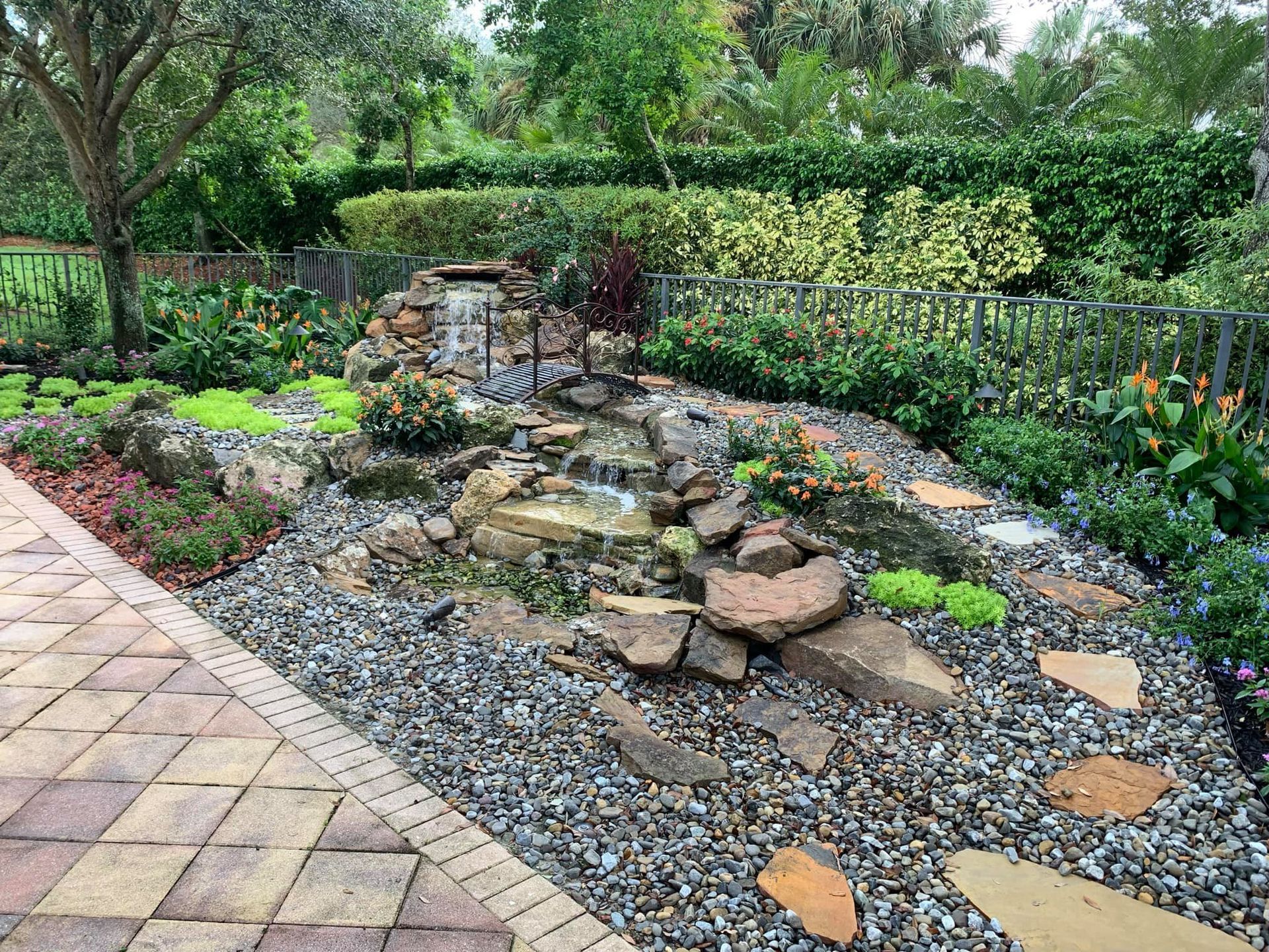 Garden pond with stepping stones, gravel, and lush green shrubs surrounded by trees.