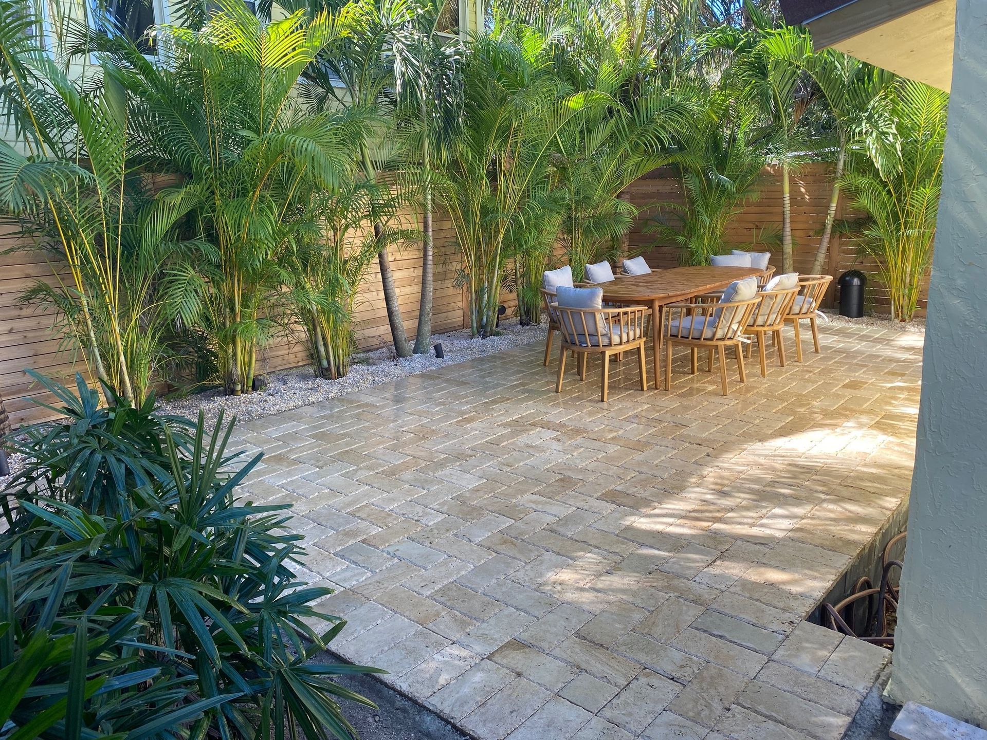 Sunny patio with stone paving, palm plants, and wooden outdoor dining table and chairs under a canopy