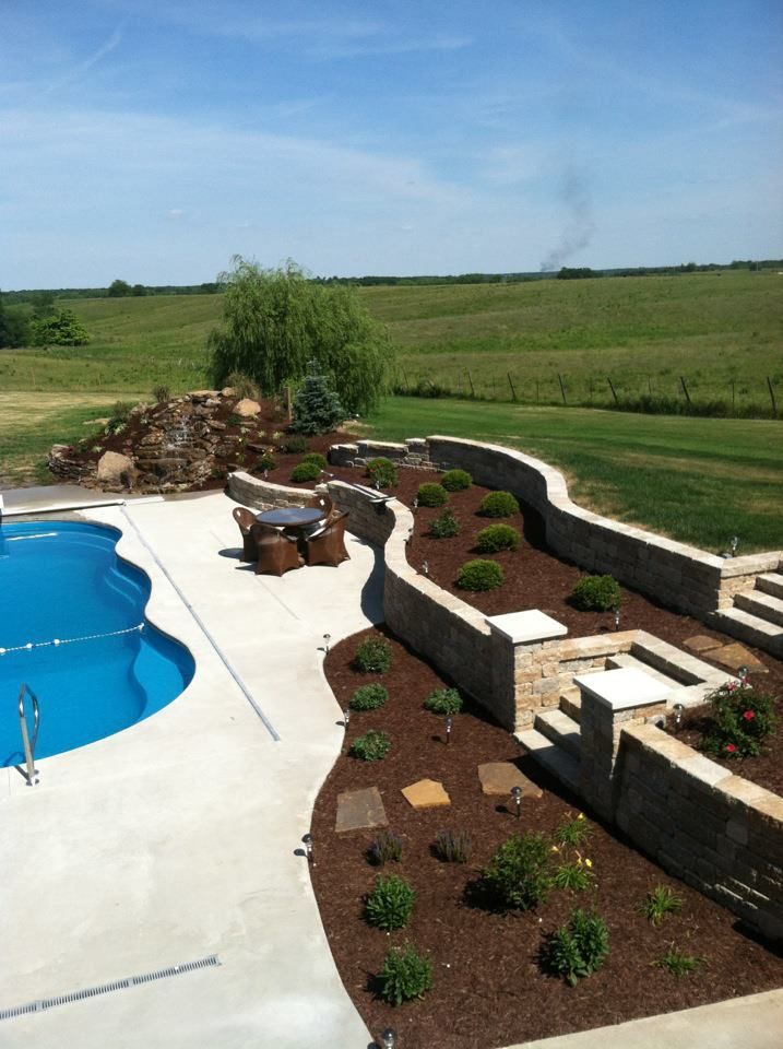 Backyard pool beside a landscaped stone retaining wall and flower beds under a blue sky