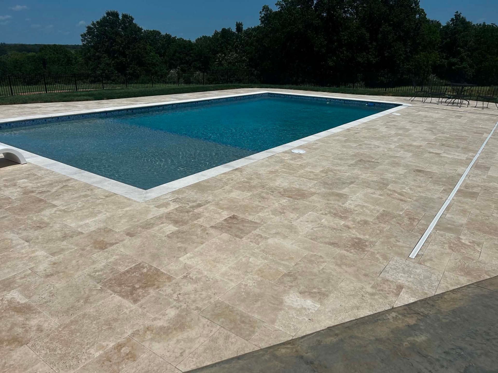 Rectangular outdoor swimming pool beside a large beige stone patio, with trees in the background
