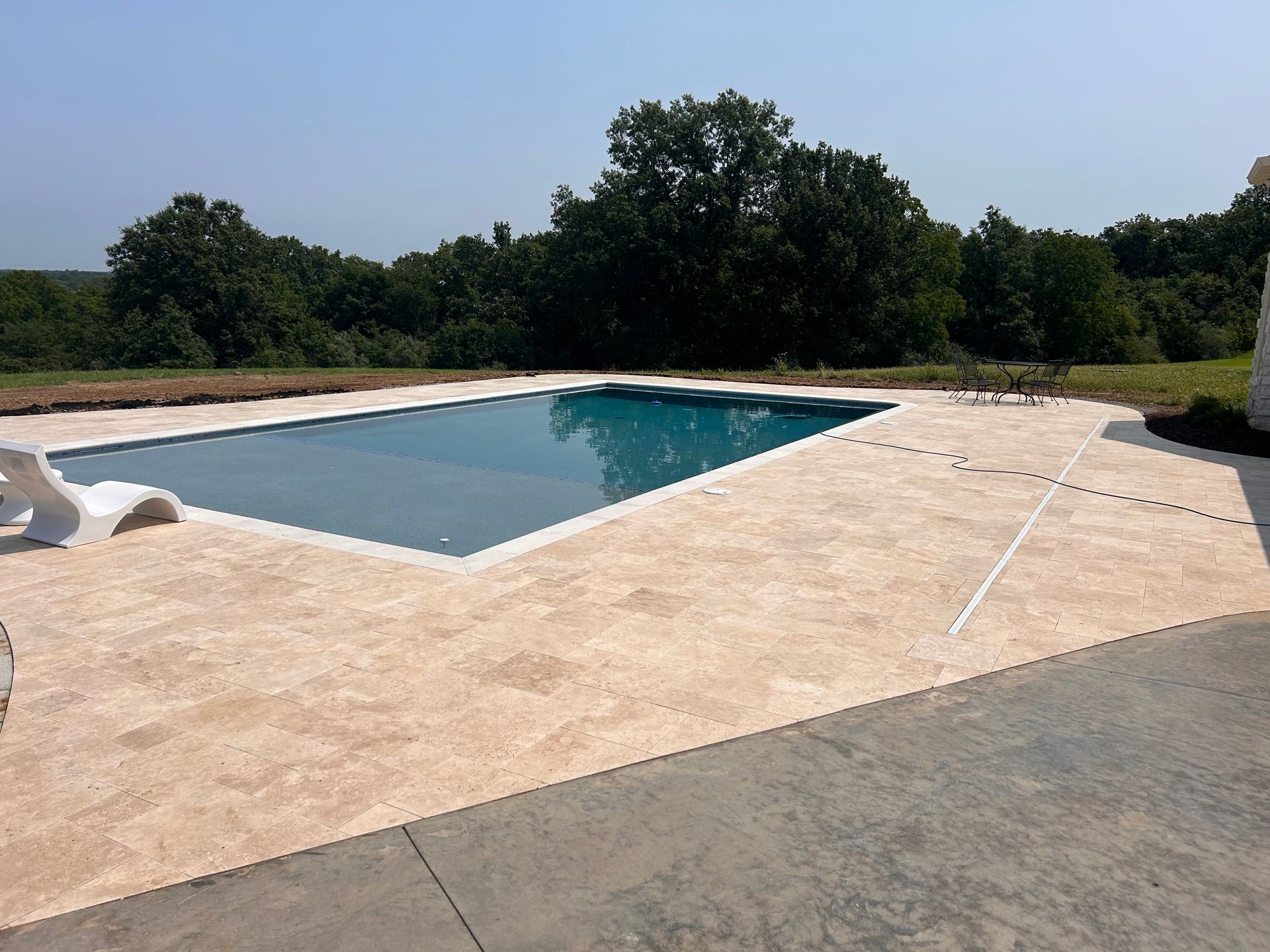 Empty backyard swimming pool with blue water and tan stone patio under a sunny sky