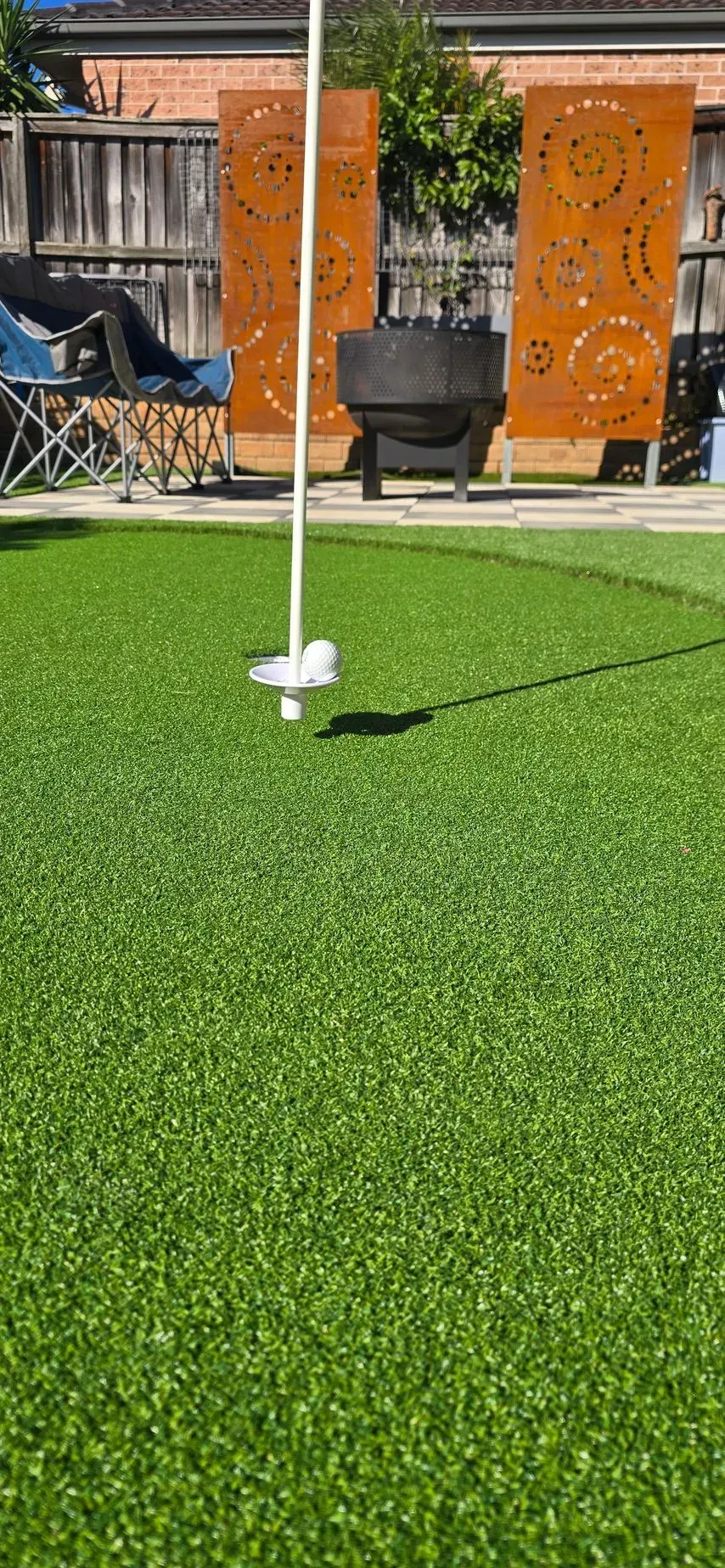 White patio umbrella base on artificial grass in a backyard with orange privacy screens and a black chair