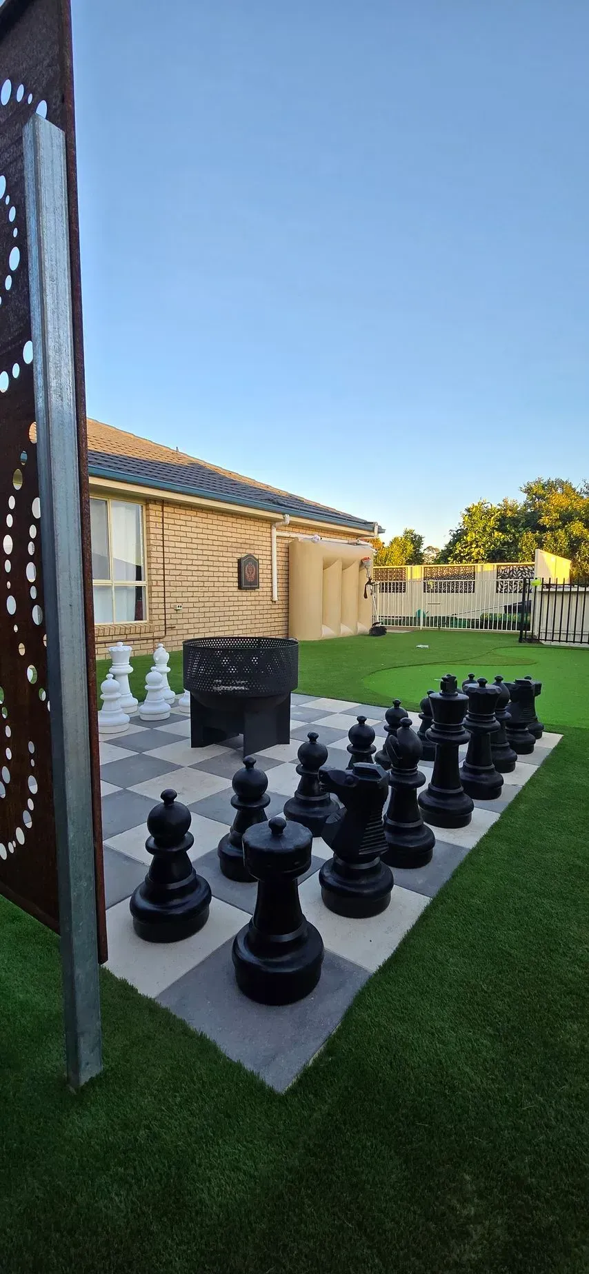 Large outdoor chess set on a patio beside a lawn and stone building under a clear blue sky.