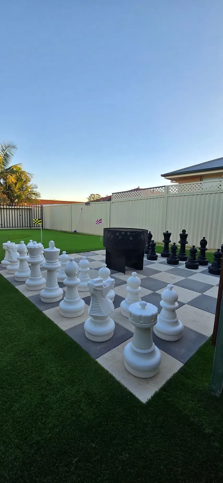 Outdoor giant chess set on artificial grass in a backyard with a white fence and blue sky