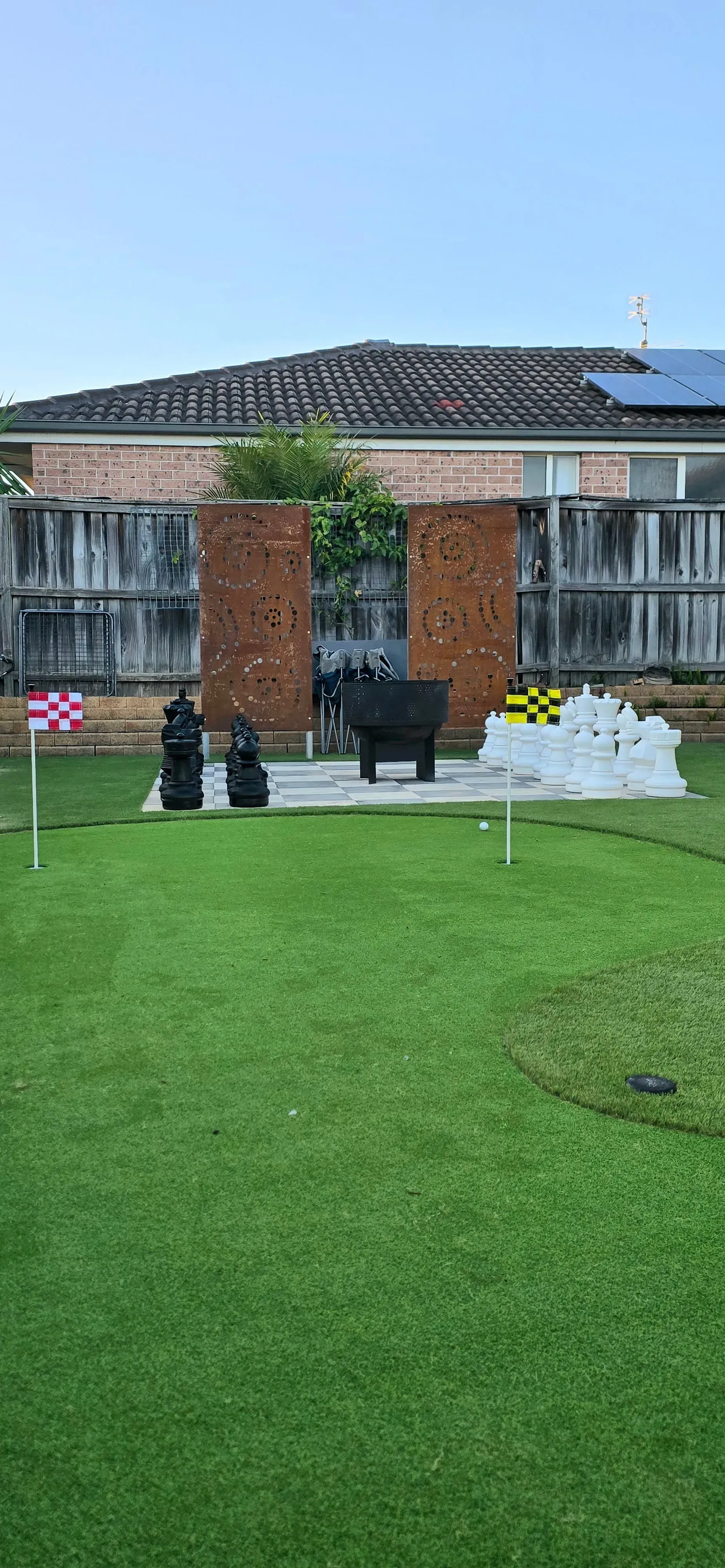 Grassy lawn with three tall chairs, two brick pillars, and a building with a long roof in the background