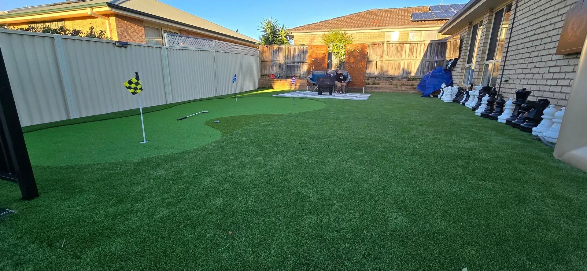 Backyard with green turf lawn, fence on the left, stone house on the right, and a small patio at the back