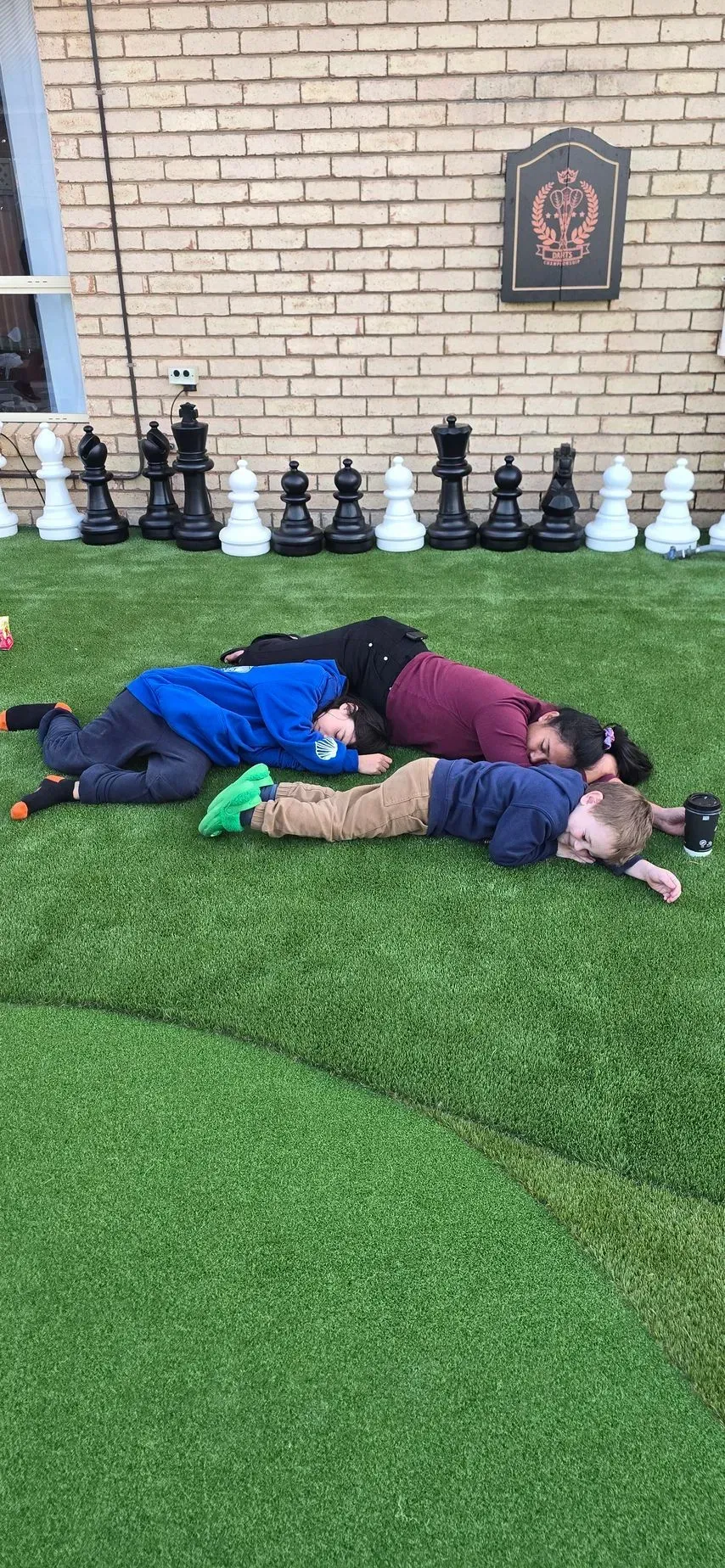 Three children lying on artificial grass beside a brick wall and a row of chess pieces