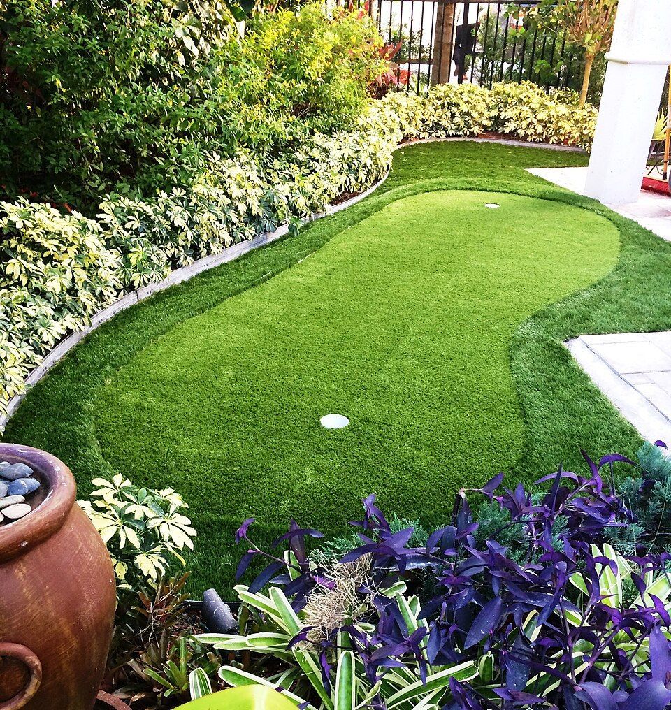 Curved artificial turf lawn bordered by lush plants and flowers in a garden walkway.