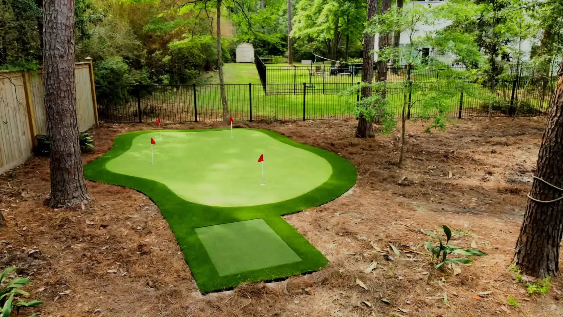 Backyard mini golf putting green with two red flags, surrounded by trees and mulch