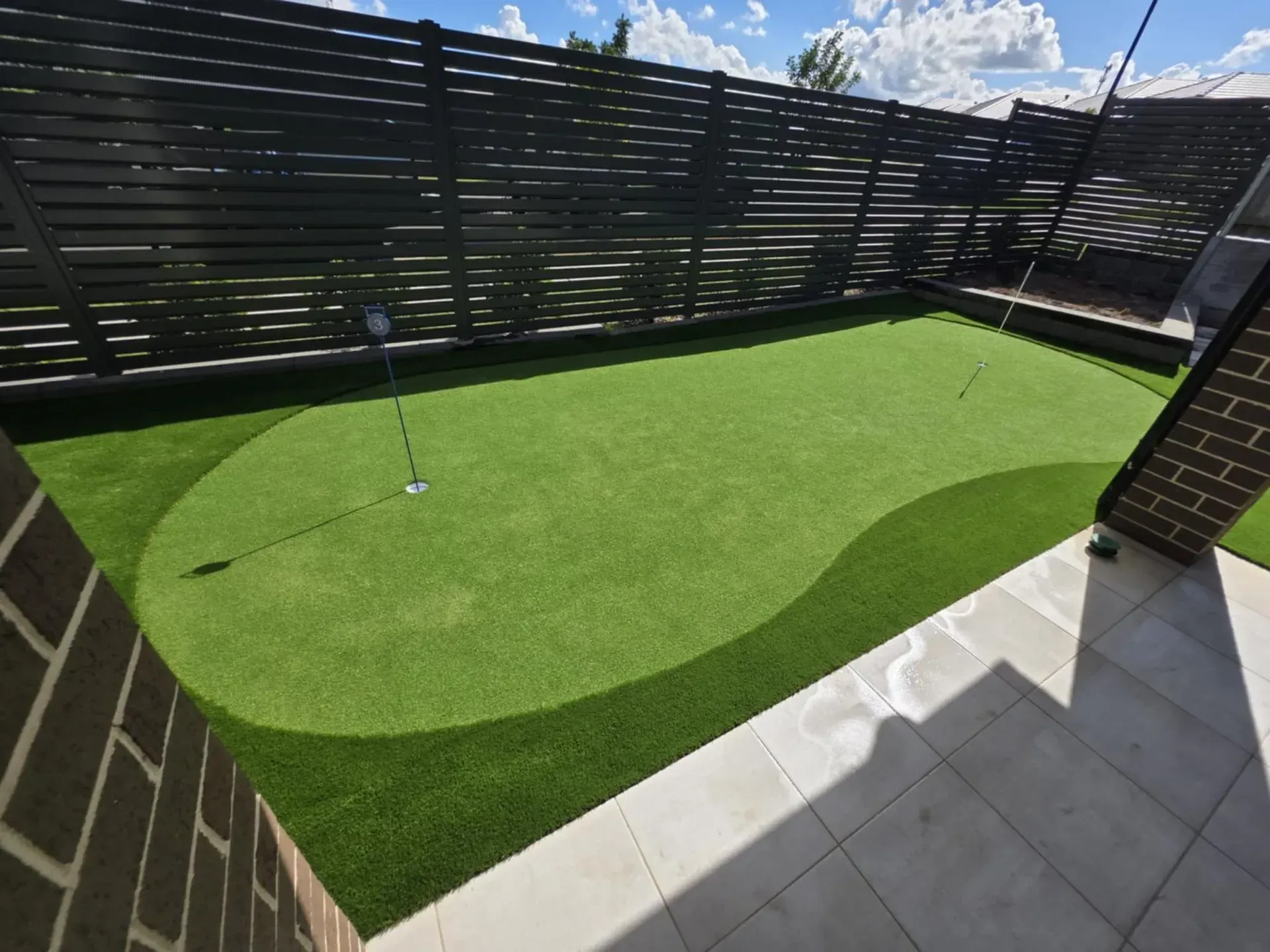 Small backyard putting green with a golf hole, surrounded by black fence and light stone patio