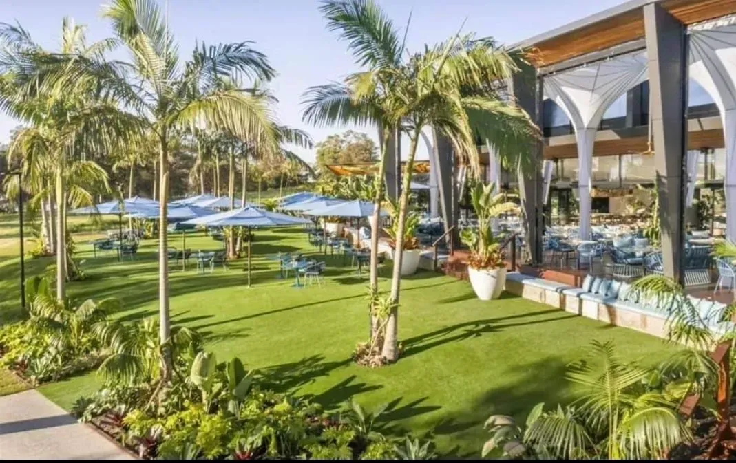 Tropical resort garden with palm trees, green lawn, blue umbrellas, and a modern poolside pavilion