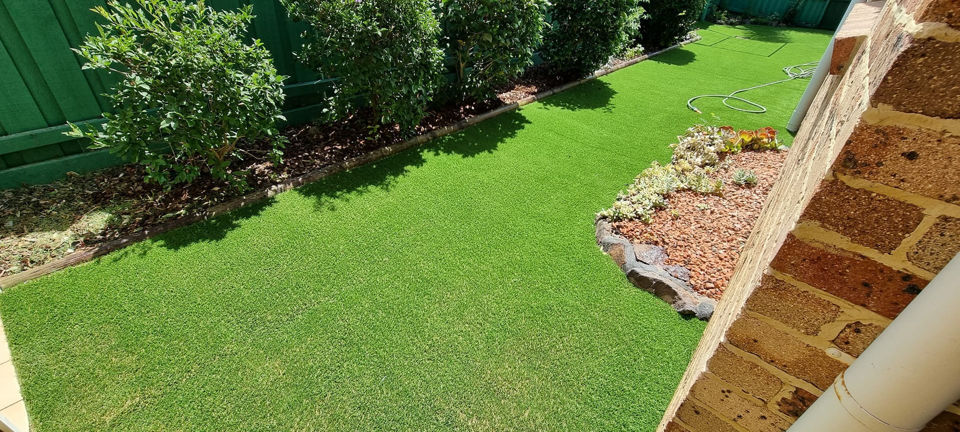 Backyard lawn beside shrubs and mulch edging next to a brick wall in sunlight