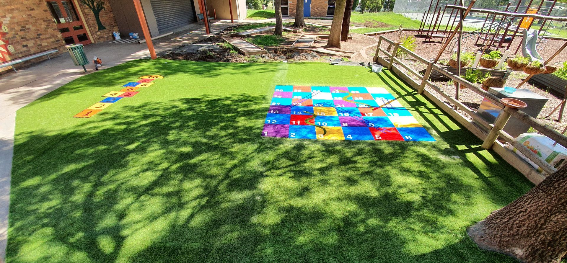 Colorful outdoor play area with green turf, multicolored mats, and benches in a sunny courtyard