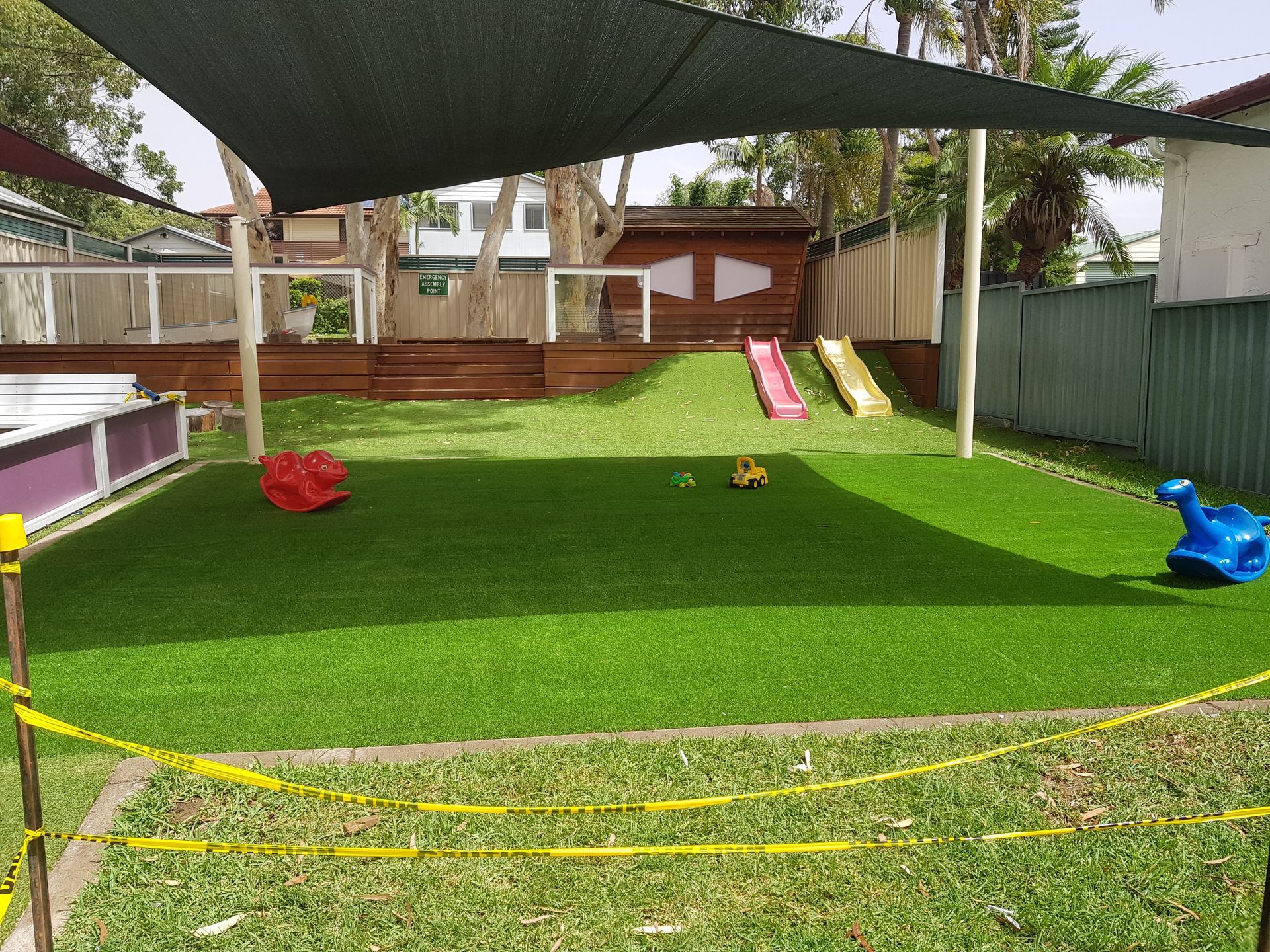 Artificial grass backyard play area with slides, a shade sail, and colorful toys