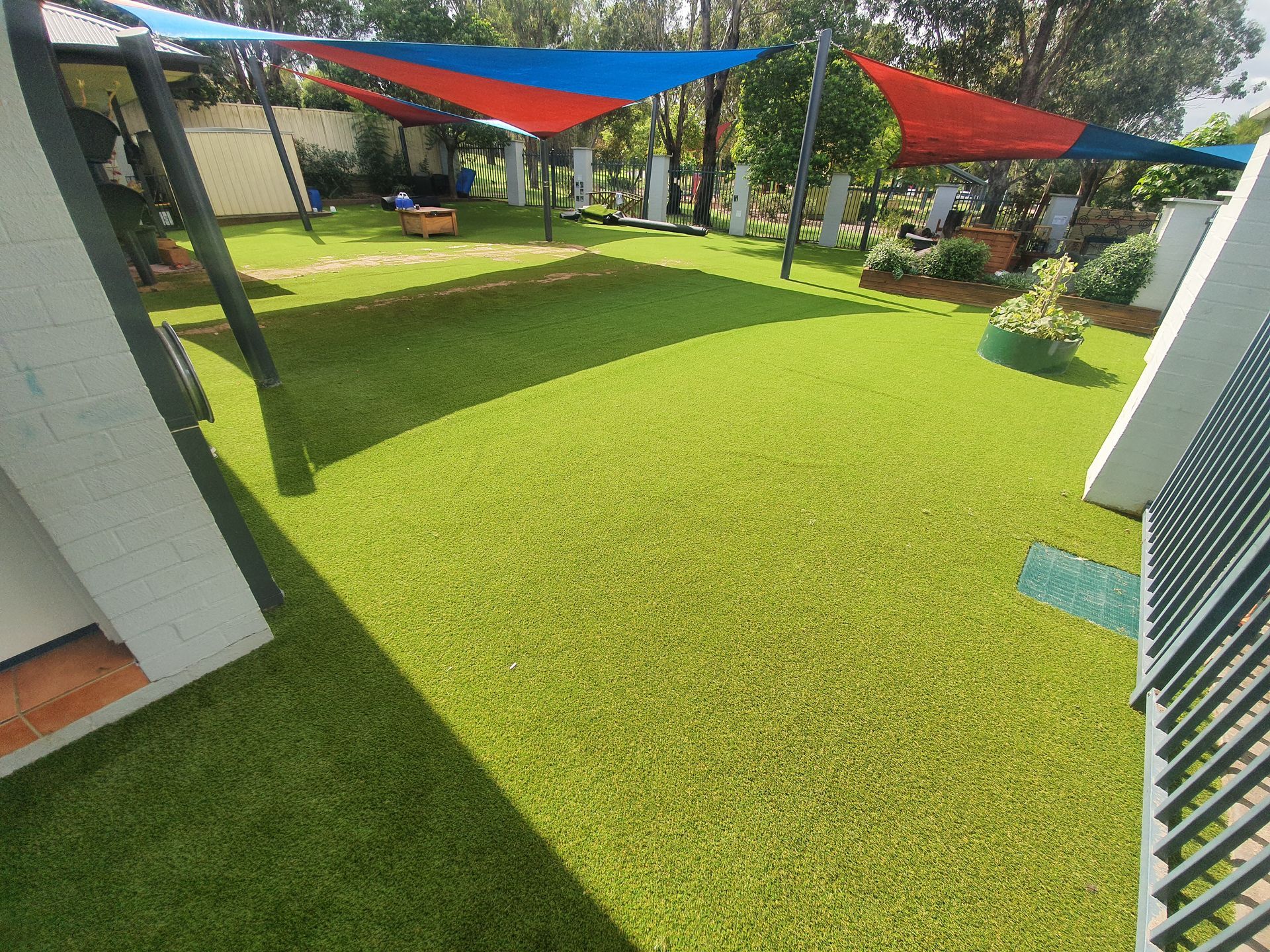 Backyard patio with green artificial grass and red and blue shade sails overhead