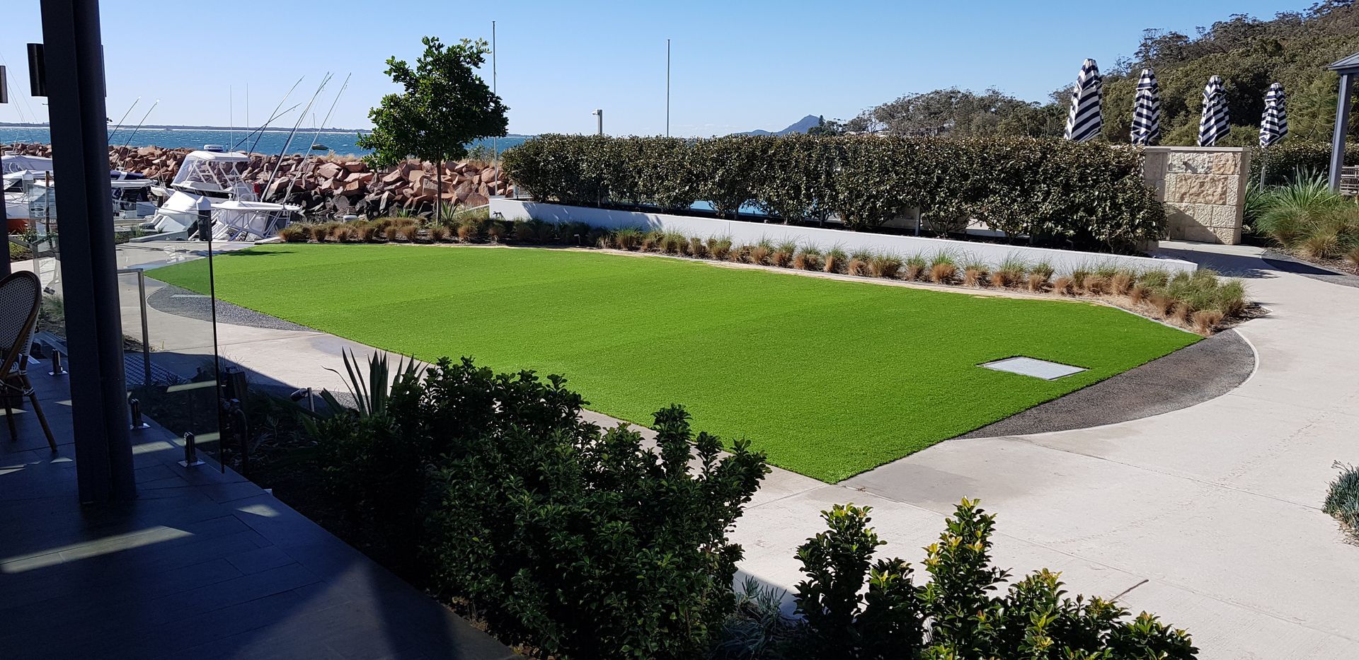 Marina walkway with green lawn, curved path, shrubs, and boats in the background on a sunny day