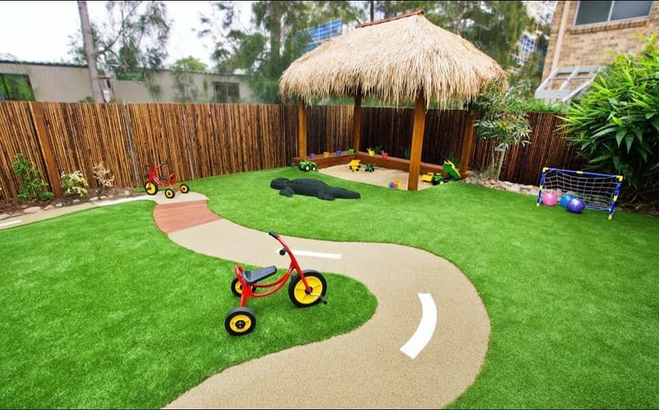 Backyard play area with curving path, red tricycle, green lawn, and thatched play hut