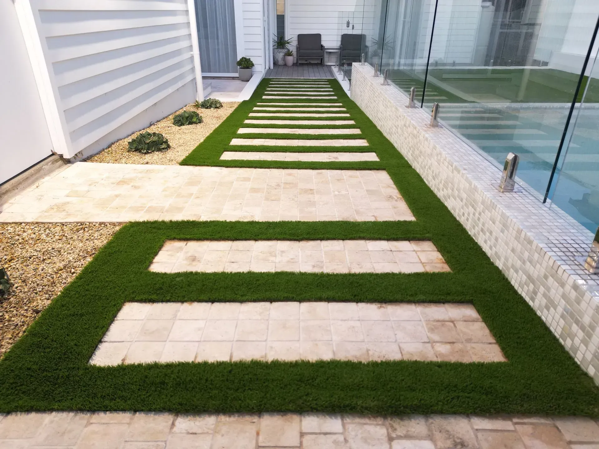 Modern backyard walkway with concrete stepping stones in artificial grass beside a glass fence and house wall