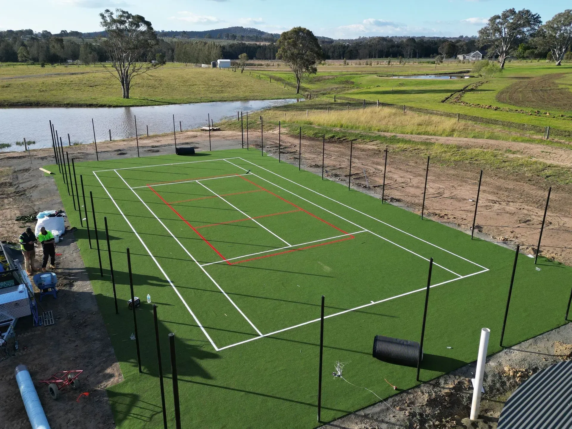 Aerial view of a fenced green sports court with white and red lines beside a pond and fields