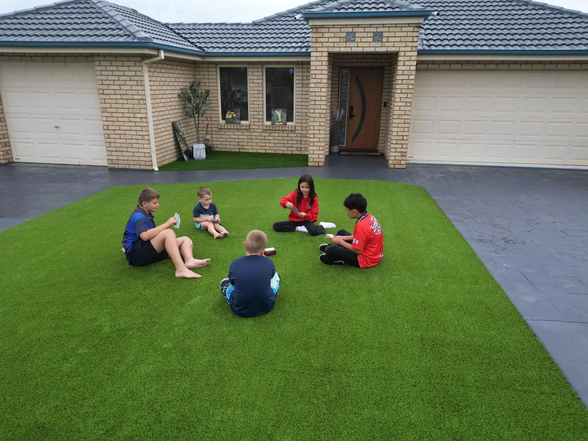 Five children sit on a green lawn in front of a suburban house and garage.