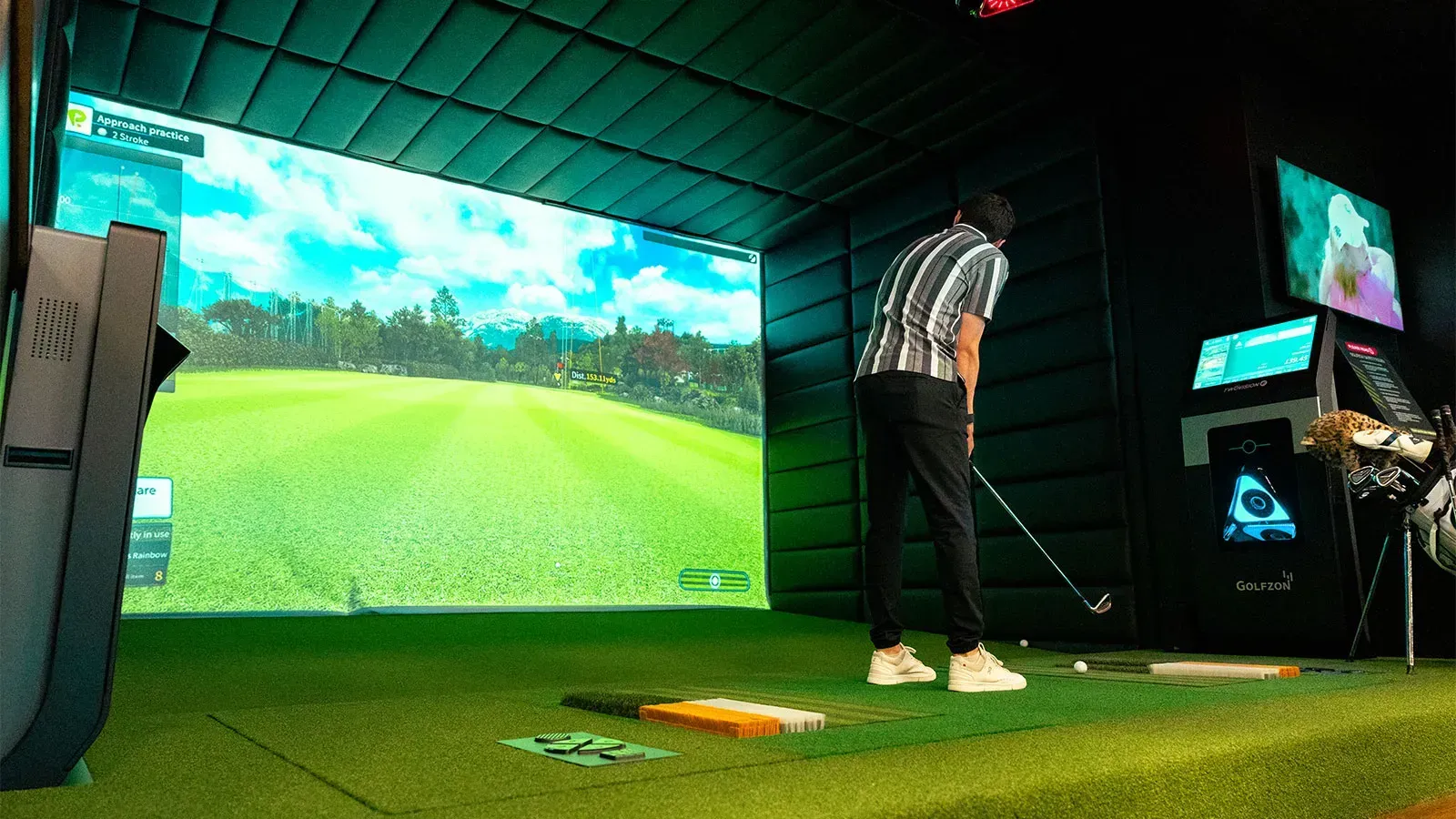 Person practicing golf indoors in a simulator room, standing on green turf facing a large virtual fairway screen.