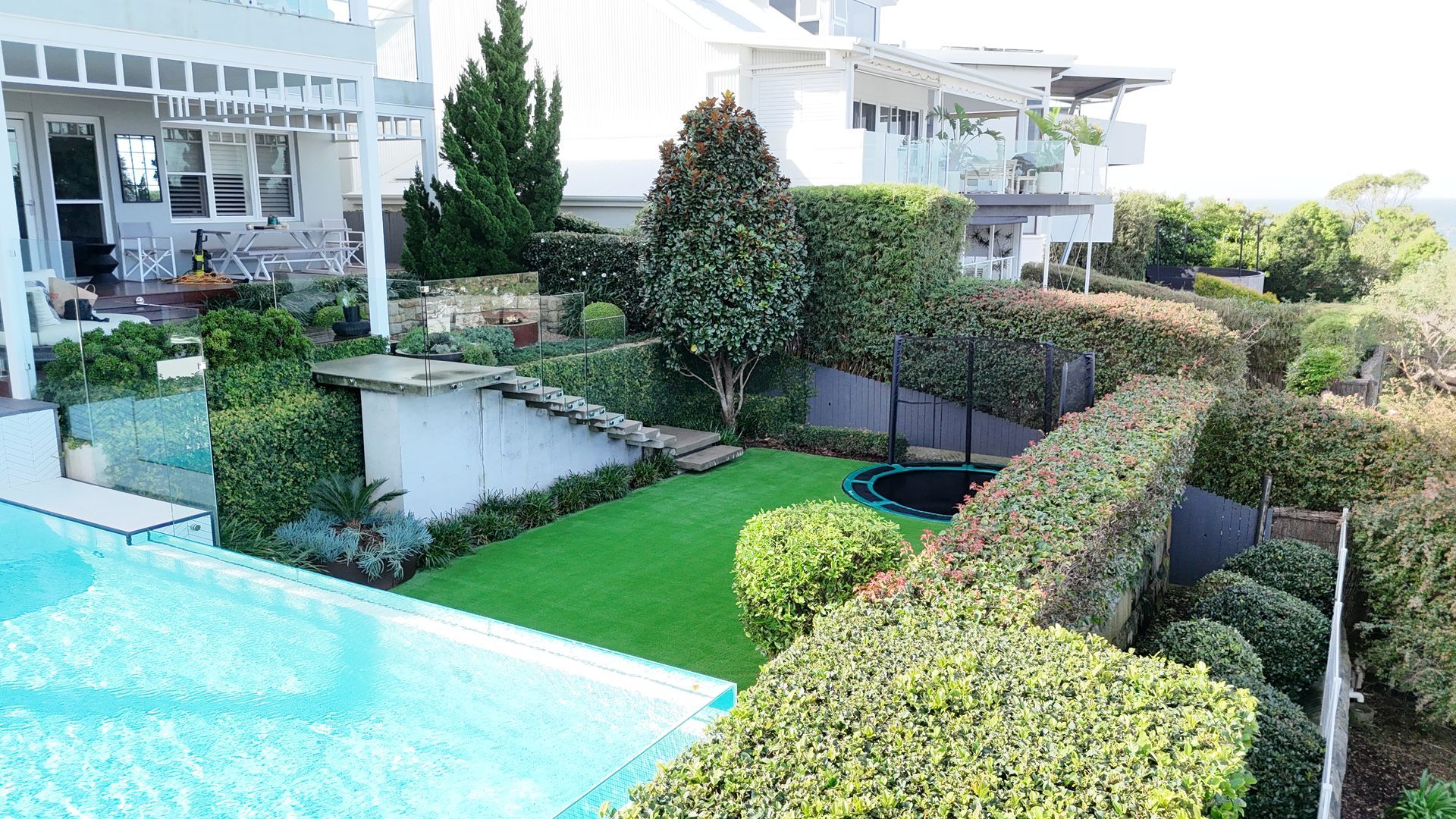 Modern backyard with turquoise pool, manicured hedges, green lawn, and white houses in the background