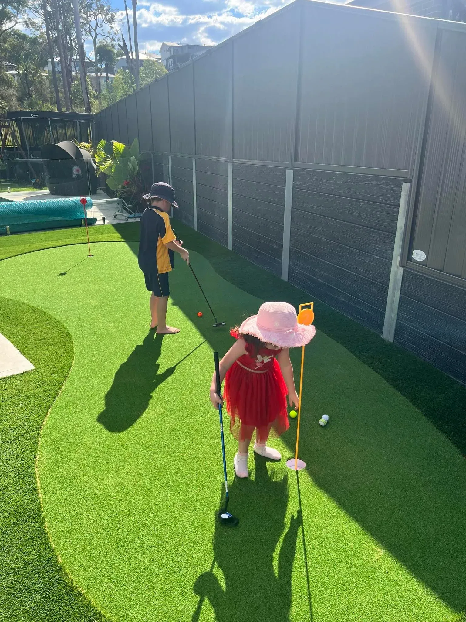 Two children play mini golf on bright green turf beside a black fence in sunny daylight