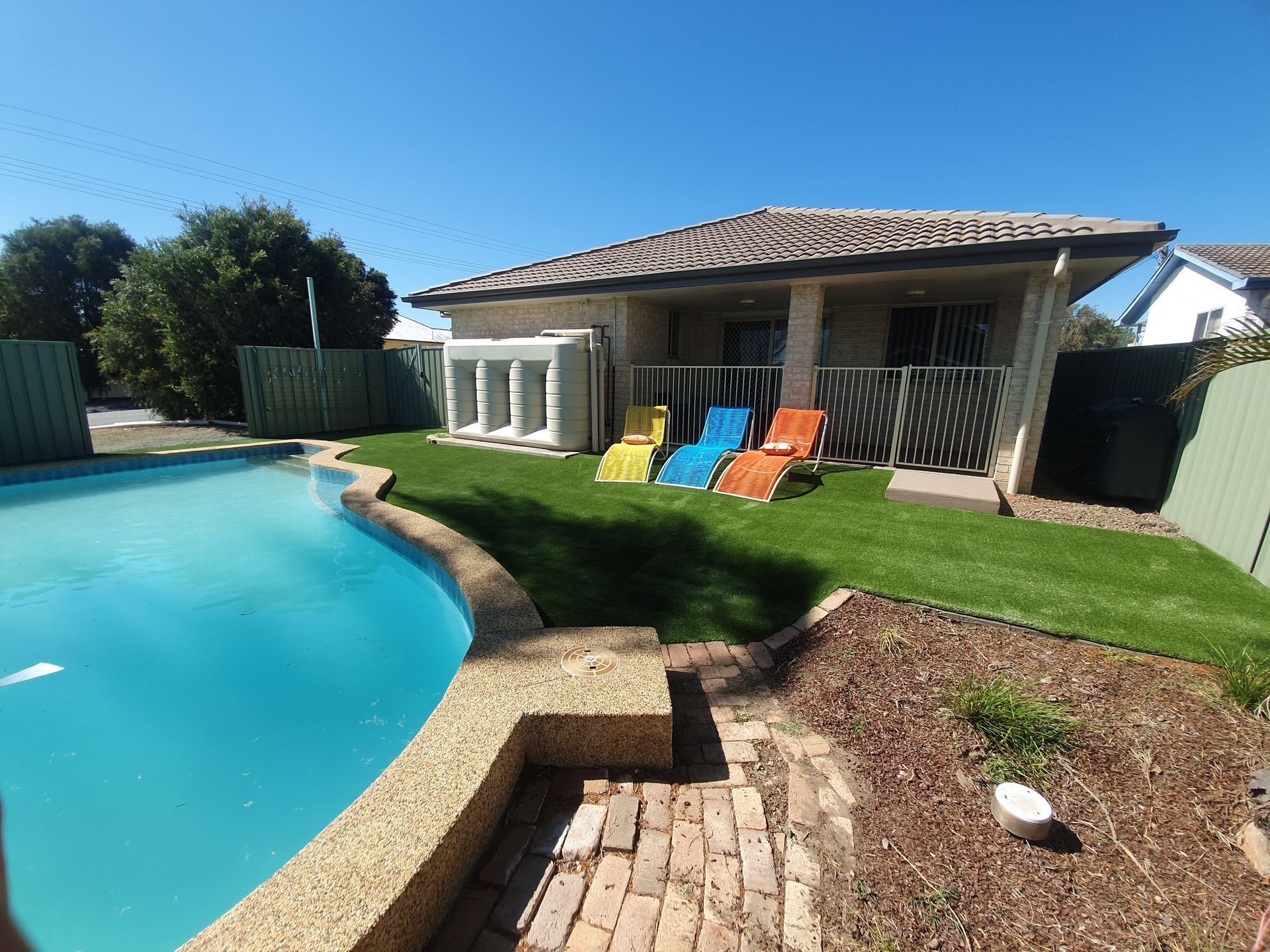 Backyard pool beside a patio with colorful lounge chairs and a tiled house under a clear blue sky