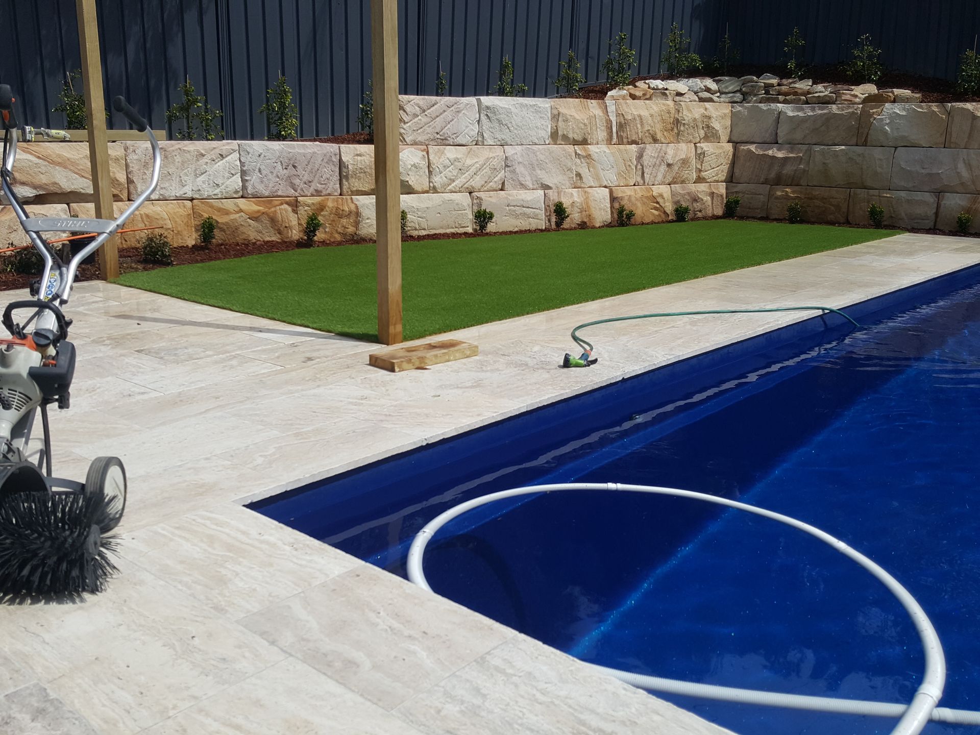 Poolside patio with a hose near a blue swimming pool and stone retaining wall
