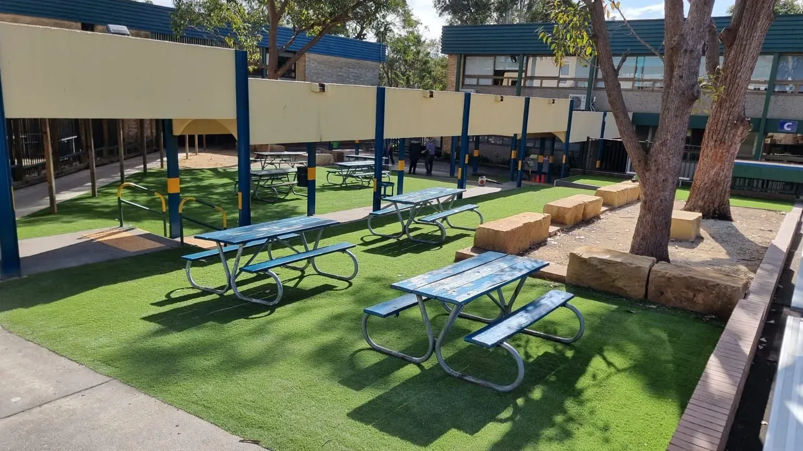 Outdoor courtyard with blue picnic tables, grass, trees, and shaded seating area