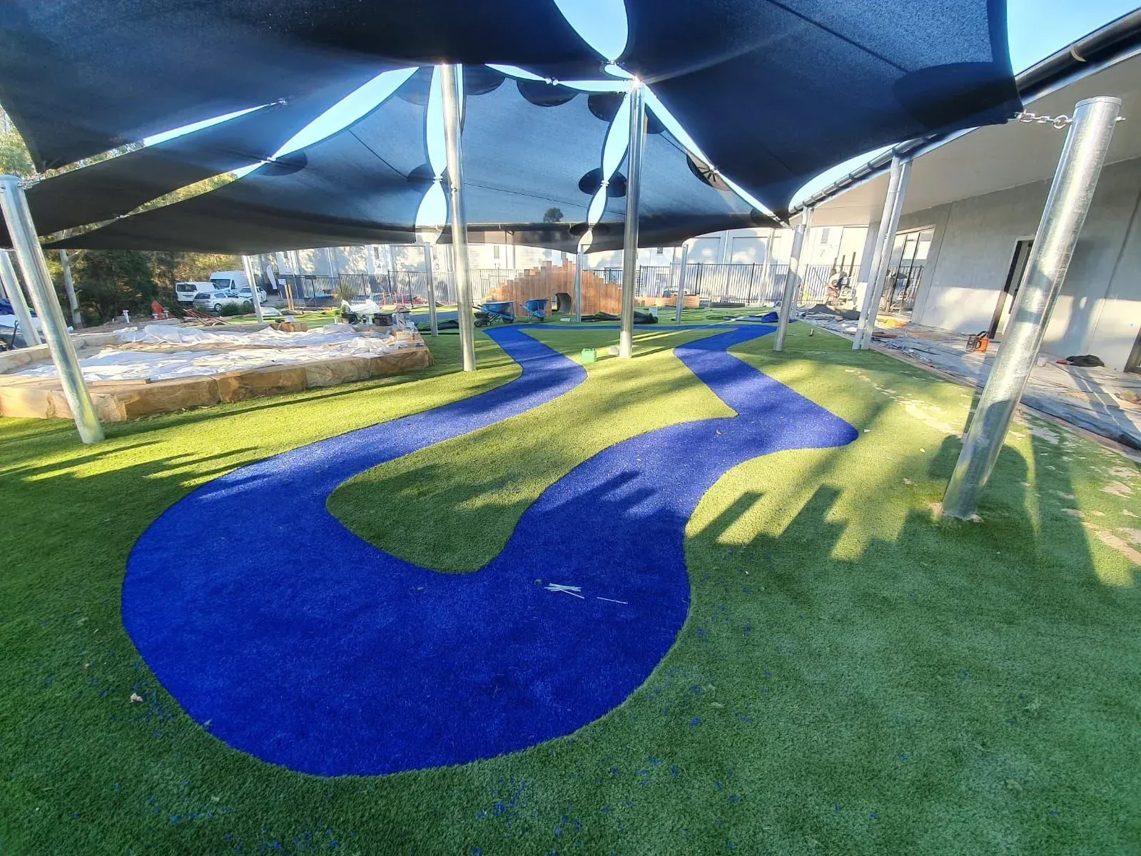 Blue wavy play path under shade sails, with green artificial turf in an outdoor playground area.
