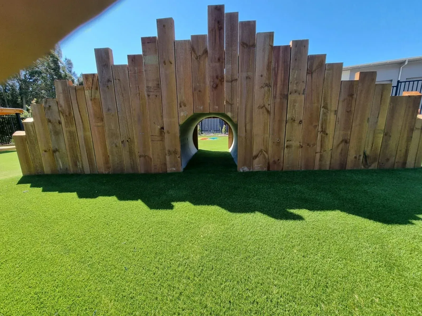 Wooden playground wall with a tunnel opening on bright green turf under a clear blue sky