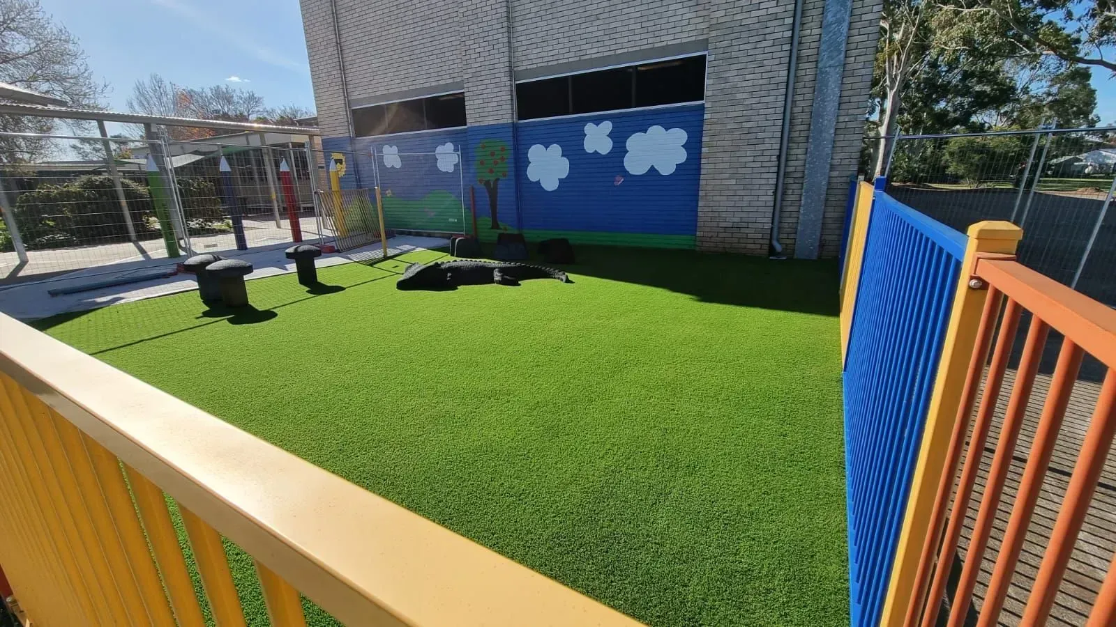 Colorful fenced playground with bright green turf, benches, and cloud mural beside a building