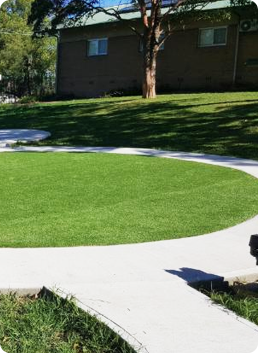 Curved concrete path around a green lawn with a tree and building in the background