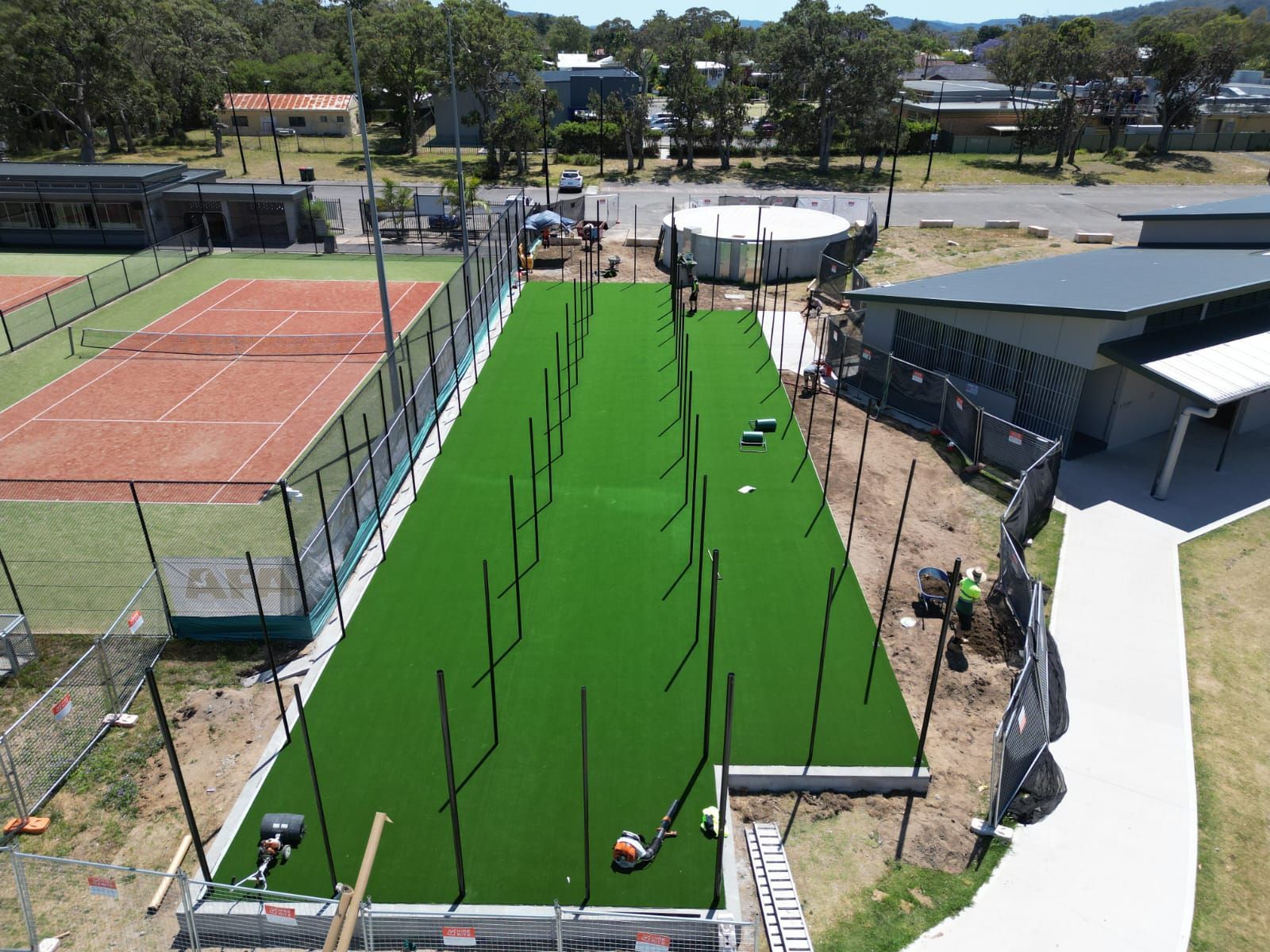 Aerial view of an outdoor sports facility with a green artificial-turf lane and nearby tennis courts.