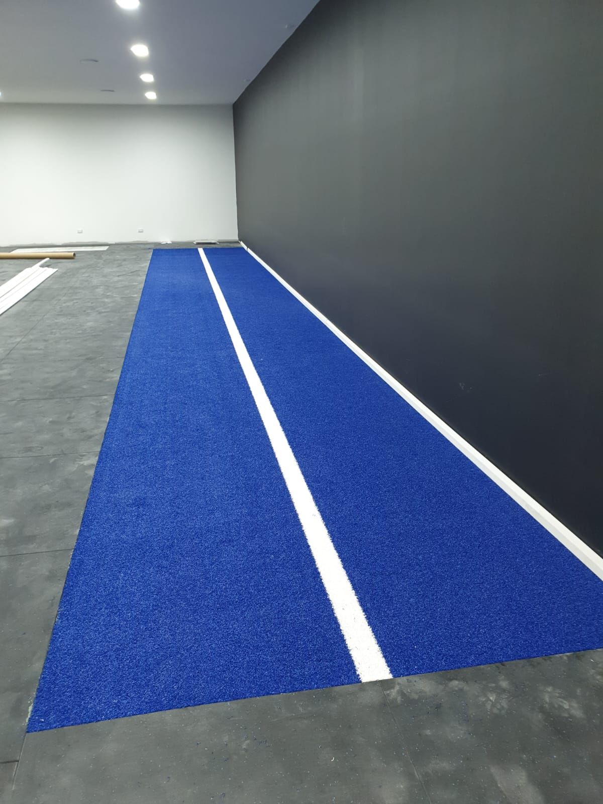 Indoor gym turf lane with blue surface and white lines beside a dark wall