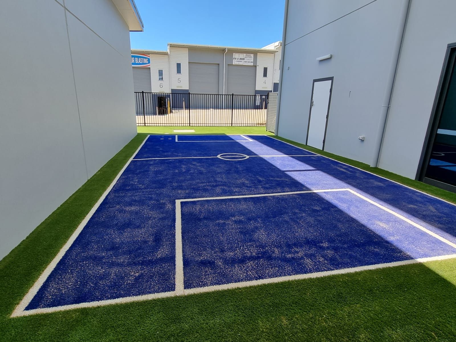 Blue pickleball court between white buildings with green turf borders and a fence at the end