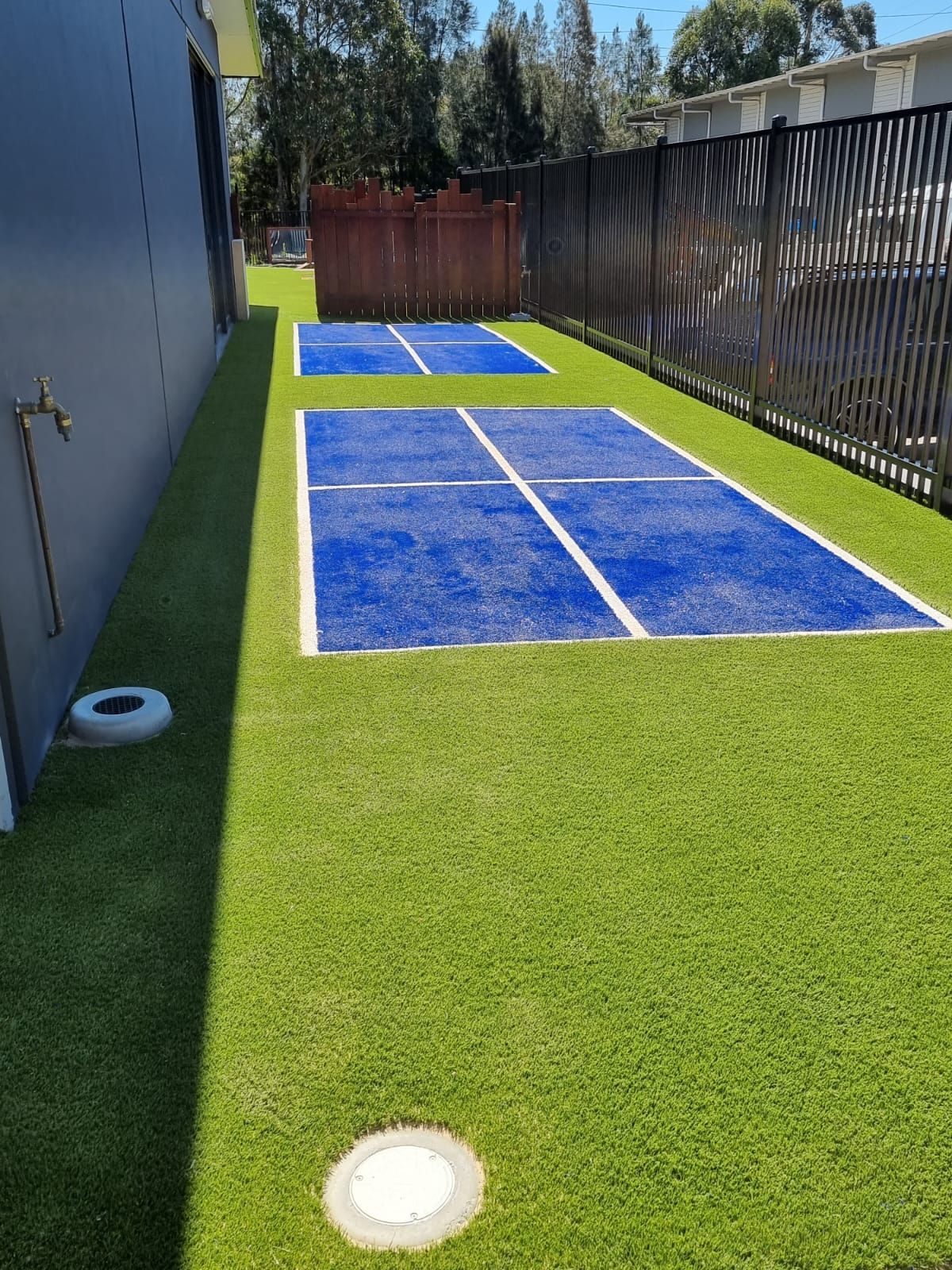 Artificial turf backyard with two blue pickleball courts between a house wall and a fence