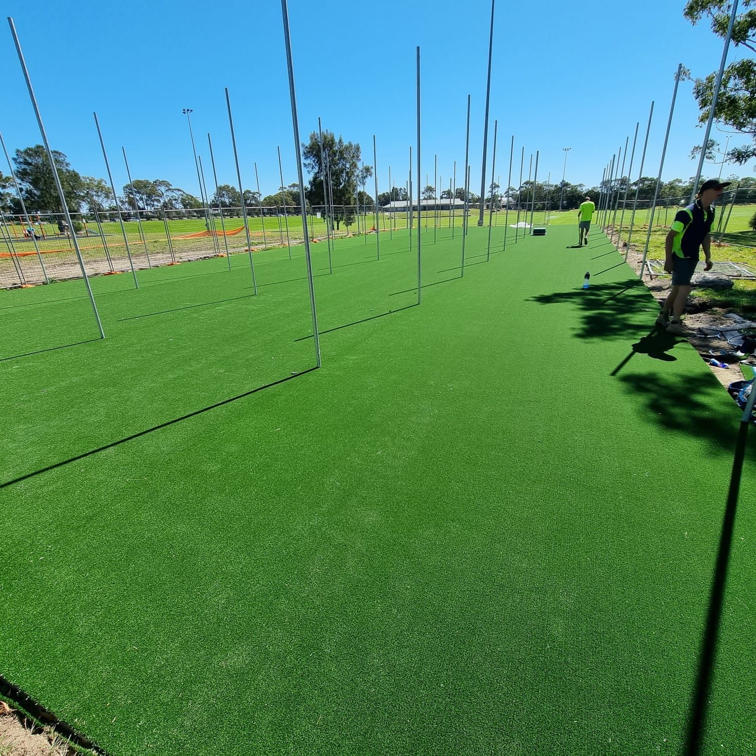 Artificial turf sports field with tall netting and a few people under a clear blue sky
