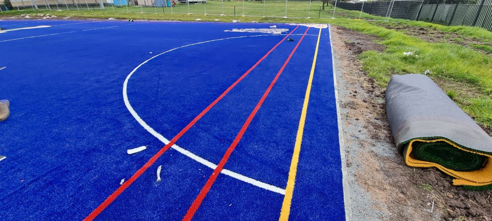 Blue sports field with white and red lines beside a gravel path and rolled turf mat