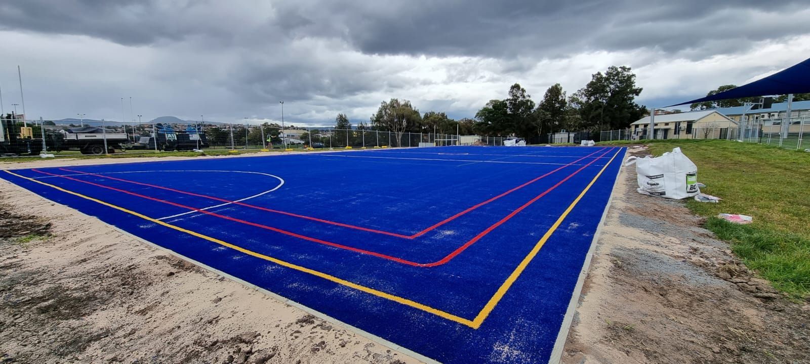 Blue sports court under cloudy sky with red and yellow lines on a dirt lot, near houses and trees