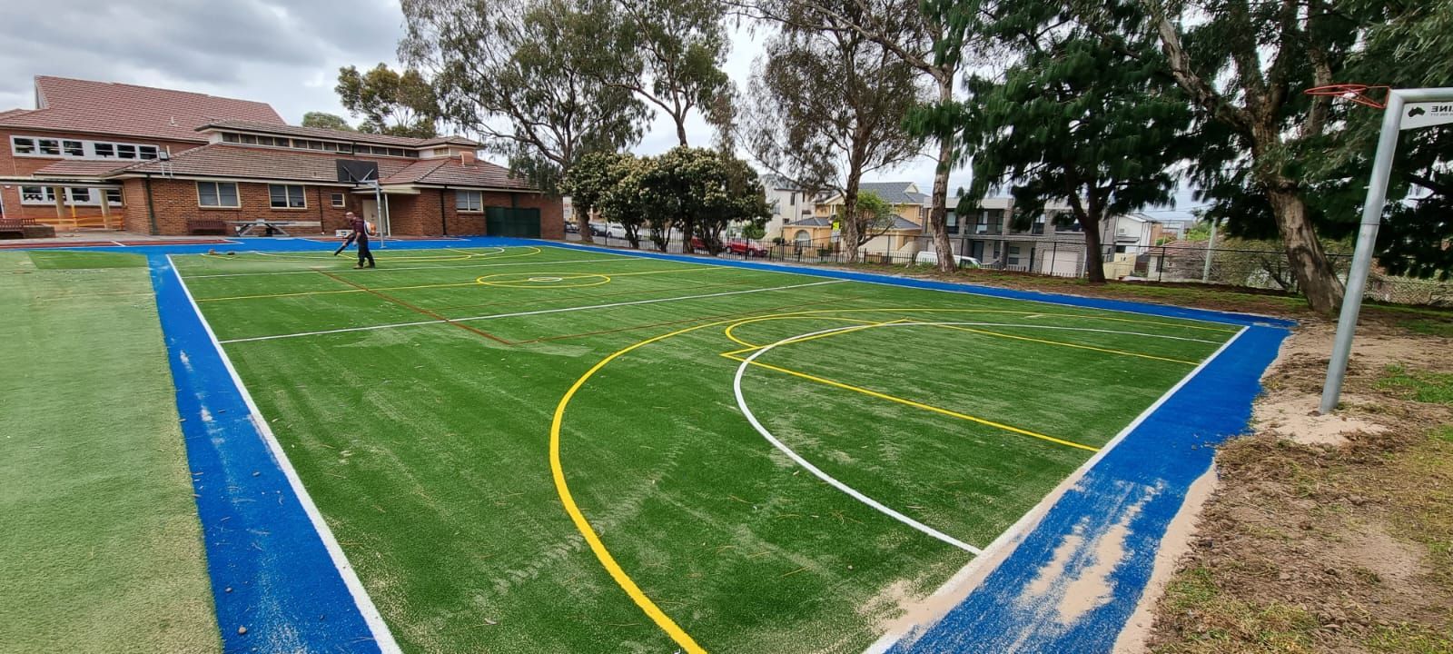 Outdoor sports field with blue sideline borders and yellow curved lines on green turf, near a brick building.
