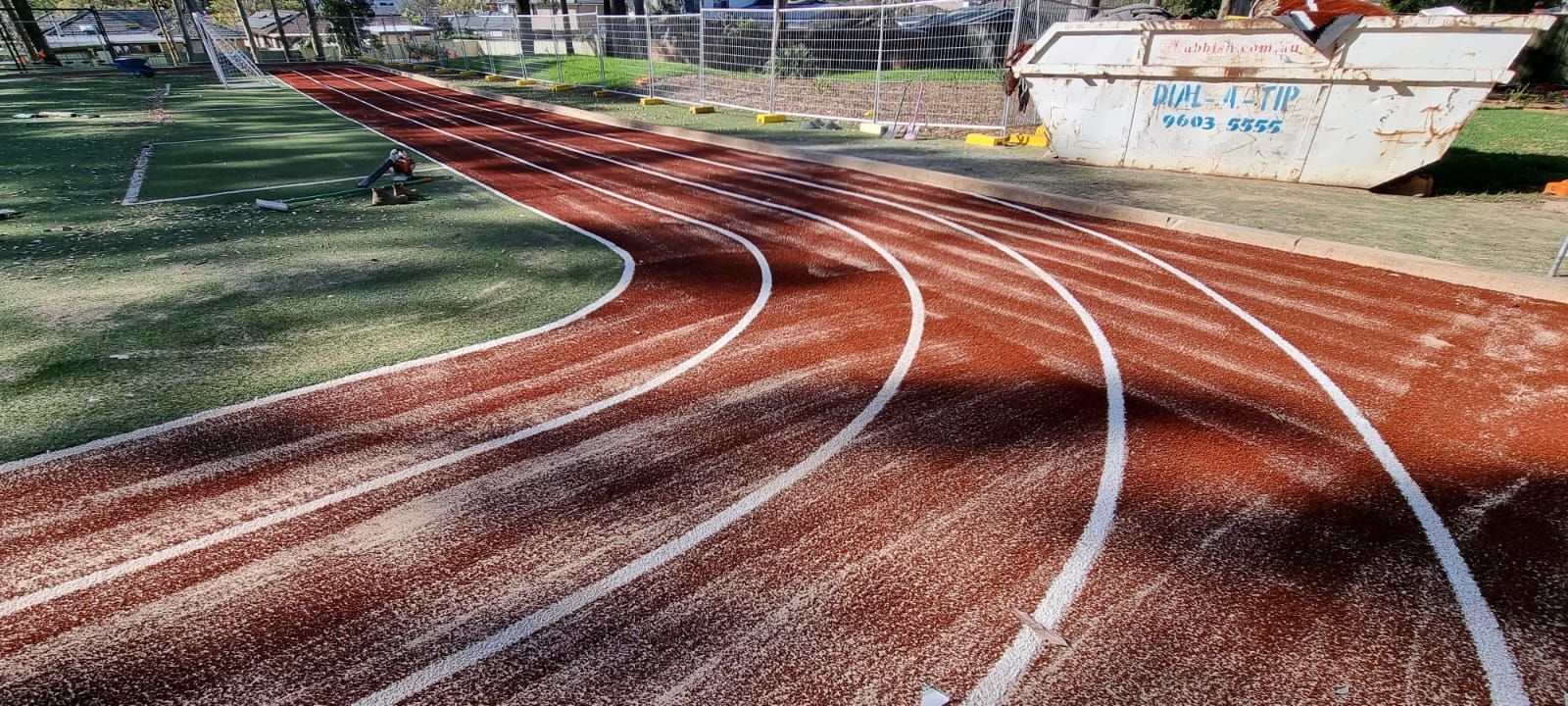 Curved red running track with white lane lines beside a grassy field and stadium seating